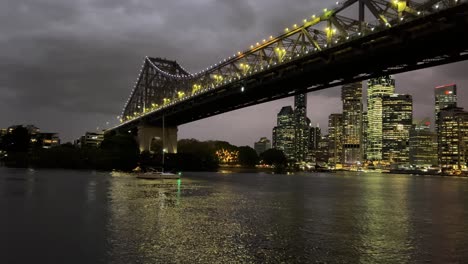 Velero-Privado-Navegando-Y-Navegando-En-El-Río-Brisbane,-Pasando-El-Puente-De-La-Historia-Hacia-El-Distrito-Central-De-Negocios-Con-Torres-De-Oficinas-Iluminadas,-Reflejo-De-Agua-Brillante-Y-Ondulada-En-Un-Día-Tormentoso