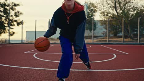 Primer-Plano-De-Una-Chica-Rubia-Con-Uniforme-Deportivo-Y-Pantalones-Azules-Maniobra-Y-Golpea-Una-Pelota-Naranja-Desde-El-Suelo-Rojo-En-Una-Cancha-De-Baloncesto-Callejera-Durante-Un-Partido-Deportivo-Por-La-Mañana.