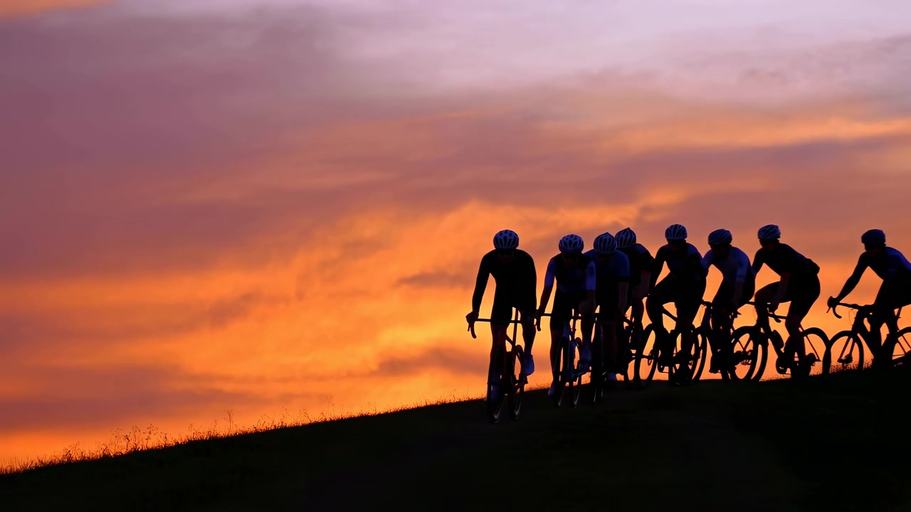 Ciclisti affiatati pedalano su bici da corsa lungo la cresta di una collina al tramonto, sagome scure contro nuvole stratificate che brillano di arancione, oro, malva, con un ampio orizzonte, un'atmosfera esterna calma ma energica