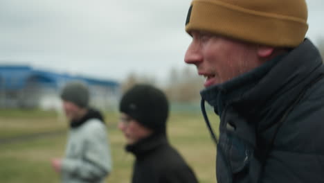close-up of three people jogging on a track near a grassy field, the coach appears to be discussing something with them, with a football post visible in the background