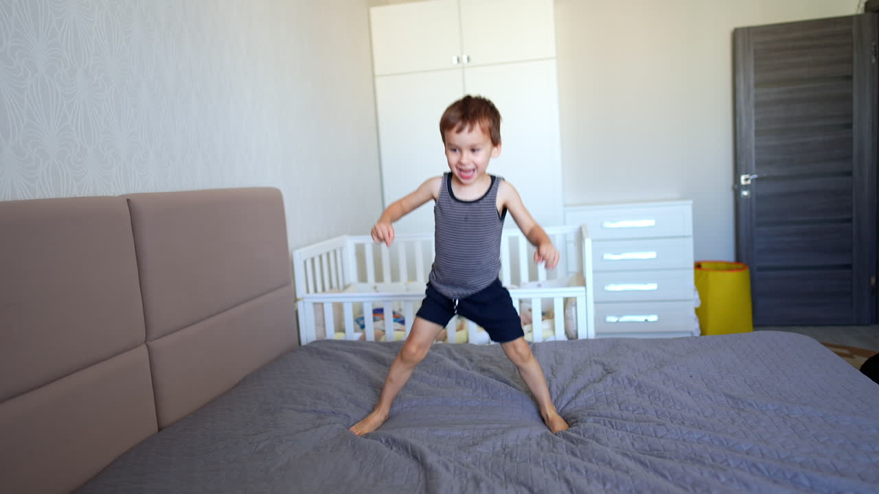 Cute baby boy in t-shirt and shorts jumps happily on the big bed. Energetic toddler having fun at home.