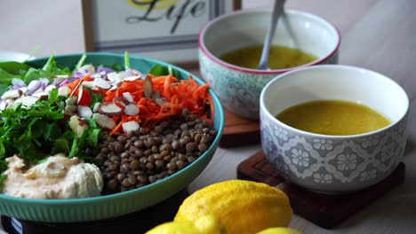 Ensalada-De-Lentejas-En-Una-Plataforma-Giratoria-Creando-Una-Ensalada-Añadiendo-Hummus-Al-Plato-Con-Rodajas-De-Espinacas-Y-Zanahorias,-Cilantro,-Cebollas,-Almendras-En-Rodajas,-Aderezo,-Limones