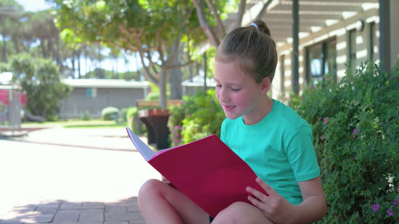 Child girl in teal shirt sitting on bricks studying red folder, camera moving closer, copy space