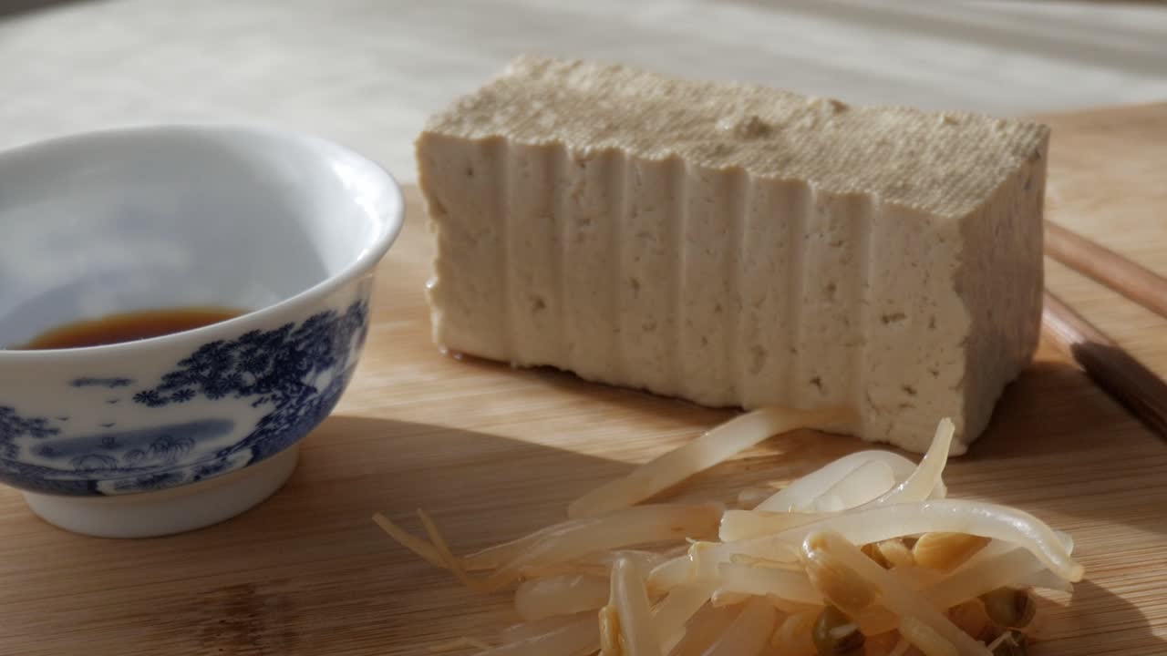 Tofu block, soy sauce in porcelain bowl and bean sprouts on wooden surface, sunny light