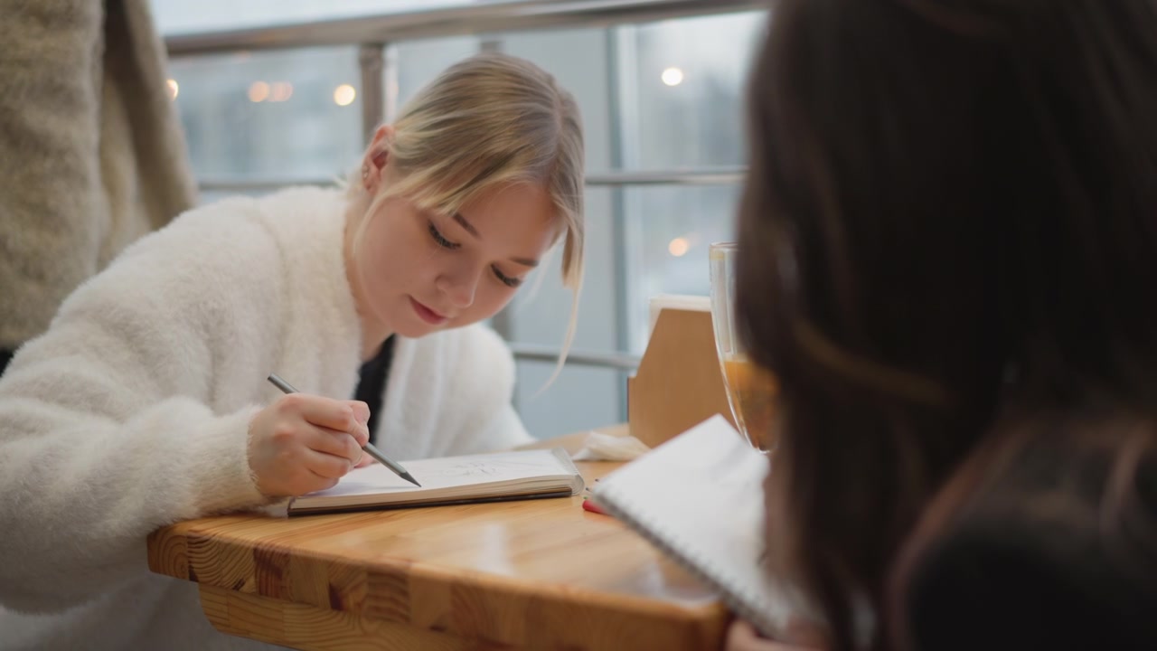 Nuori nainen nojautuu eteenpäin ja kirjoittaa muistikirjaan puisella kahvilapöydällä, kynä koskettaa paperia vakaalla kädenliikkeellä, rento asento, pehmeä päivänvalo, lämmin sisustus ja rauhallinen, tuottelias tunnelma