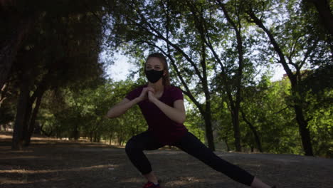 young woman using a face mask doing side lunges in a park, wide shot