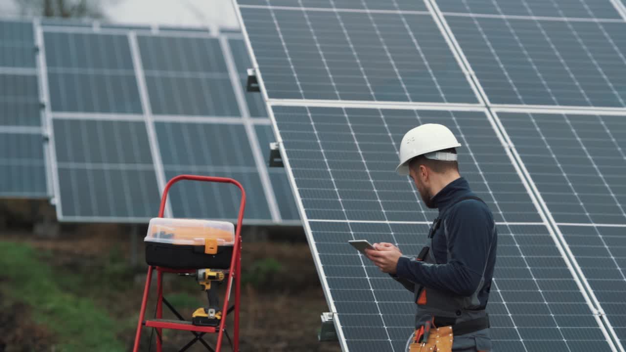 A professional man in a special electrician's uniform conducts a technical inspection of the solar station and records everything on a tablet