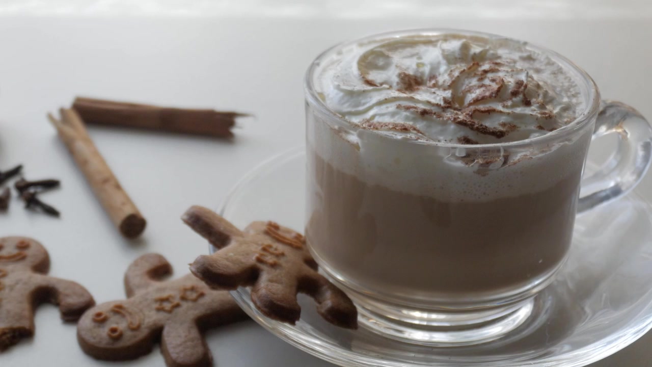 Zoom indietro su una tazza di vetro trasparente con caffè o cacao con panna, cannella, zucca, noci, omino di pan di zenzero e chiodi di garofano. Sapori natalizi.