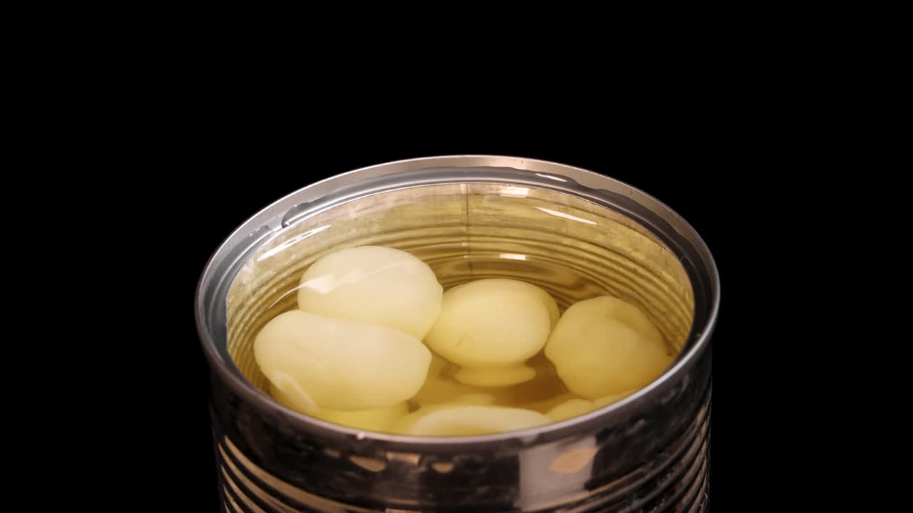Chopsticks grasp a peeled longan fruit from a can of syrup, isolated on a black background with even lighting and a static overhead camera angle