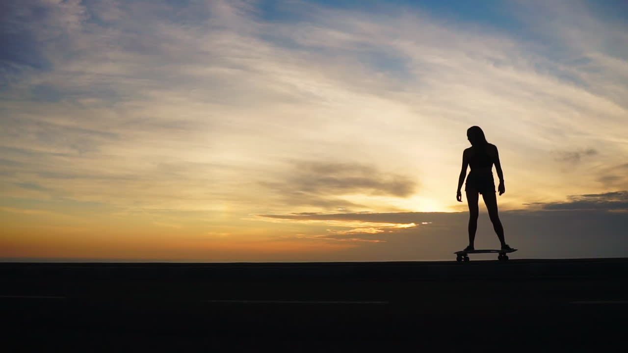 Slow-motion footage captures the essence of a woman skateboarding on a road at sunset, with mountains and a picturesque sky in view. She's attired in shorts