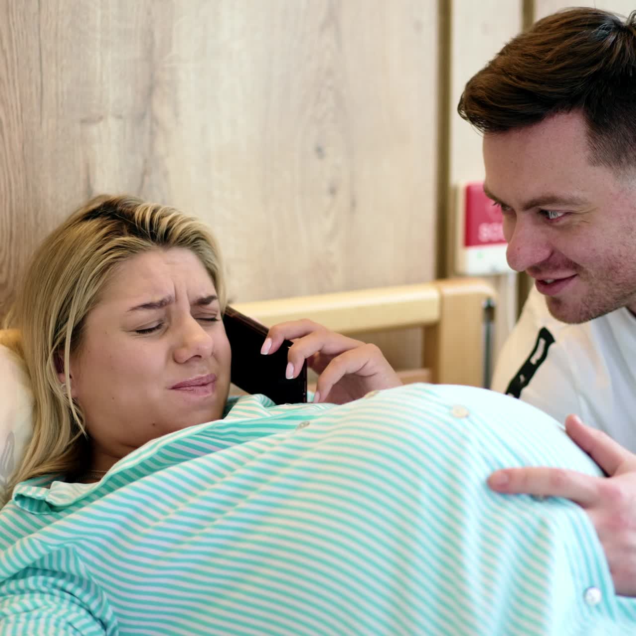 Pregnant woman lies in a bed feeling pain from contractions. Husband tries to help his wife and relief her pain