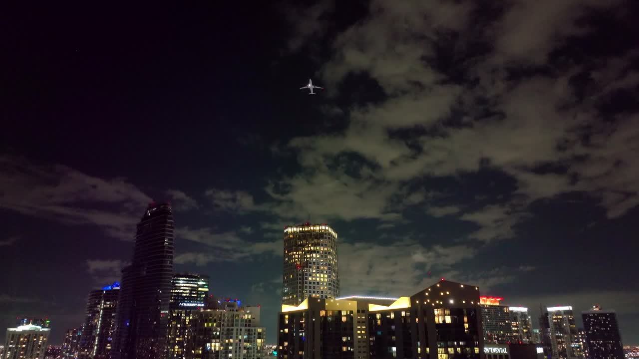 An airplane glides above Miami's illuminated downtown skyline in the evening, enhancing the city's vibrant energy