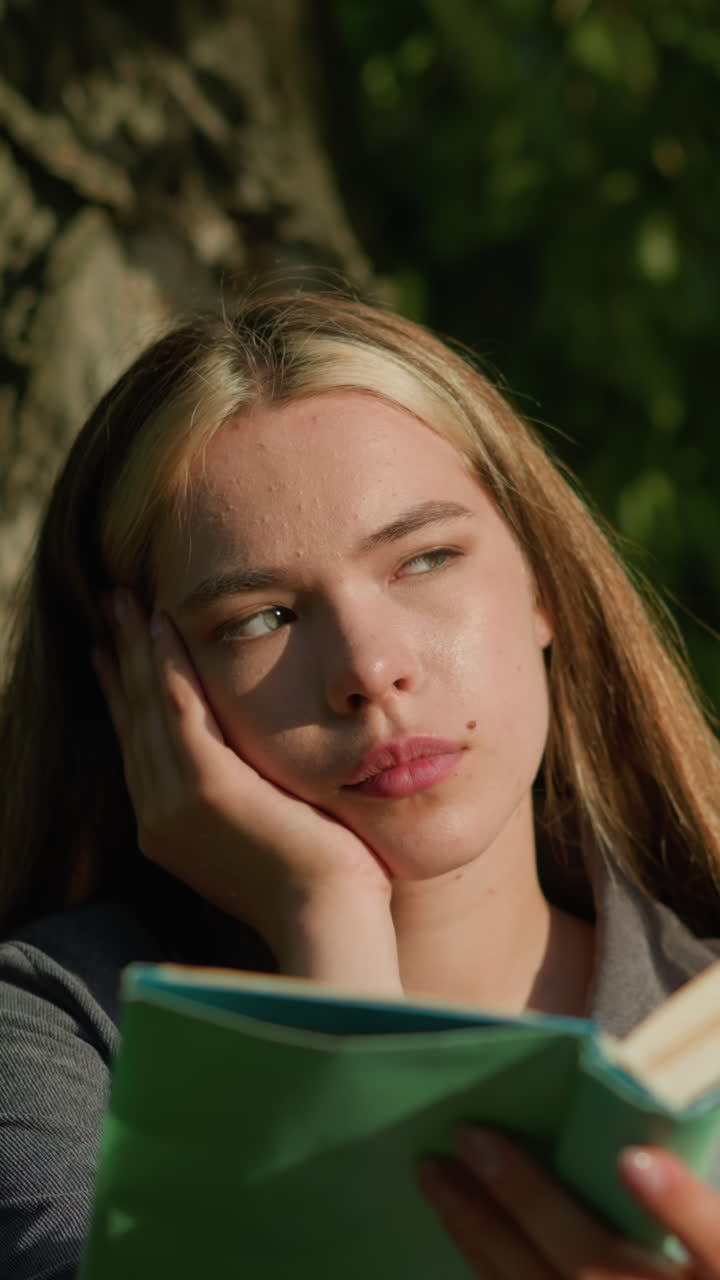 dama sentada bajo un árbol con la cabeza apoyada en su mano izquierda, leyendo un libro, las páginas volviendo en el viento, mira hacia la distancia con aspecto de mal humor, la luz del sol ilumina su rostro, el fondo suavemente borroso