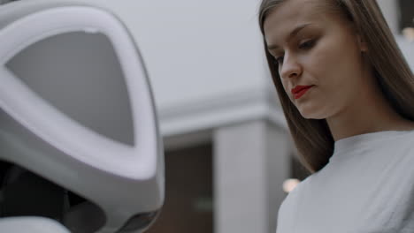 Smiling-girl-in-white-t-shirt-is-in-contact-with-humanoid.-Cyborg-helps-the-girl-answering-questions.-Useful-robot-assistant-in-the-office-and-shopping-center