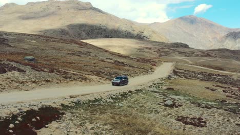 Drone-Aéreo-De-Un-Todoterreno-Rojo-Conduciendo-Lentamente-Por-Una-Colina-En-Un-Camino-De-Tierra-En-La-Llanura-Alpina-De-Gran-Altitud-Y-Las-Montañas-Del-Parque-Nacional-Deosai-Ubicado-Entre-Skardu-Y-El-Valle-De-Astore-En-Pakistán