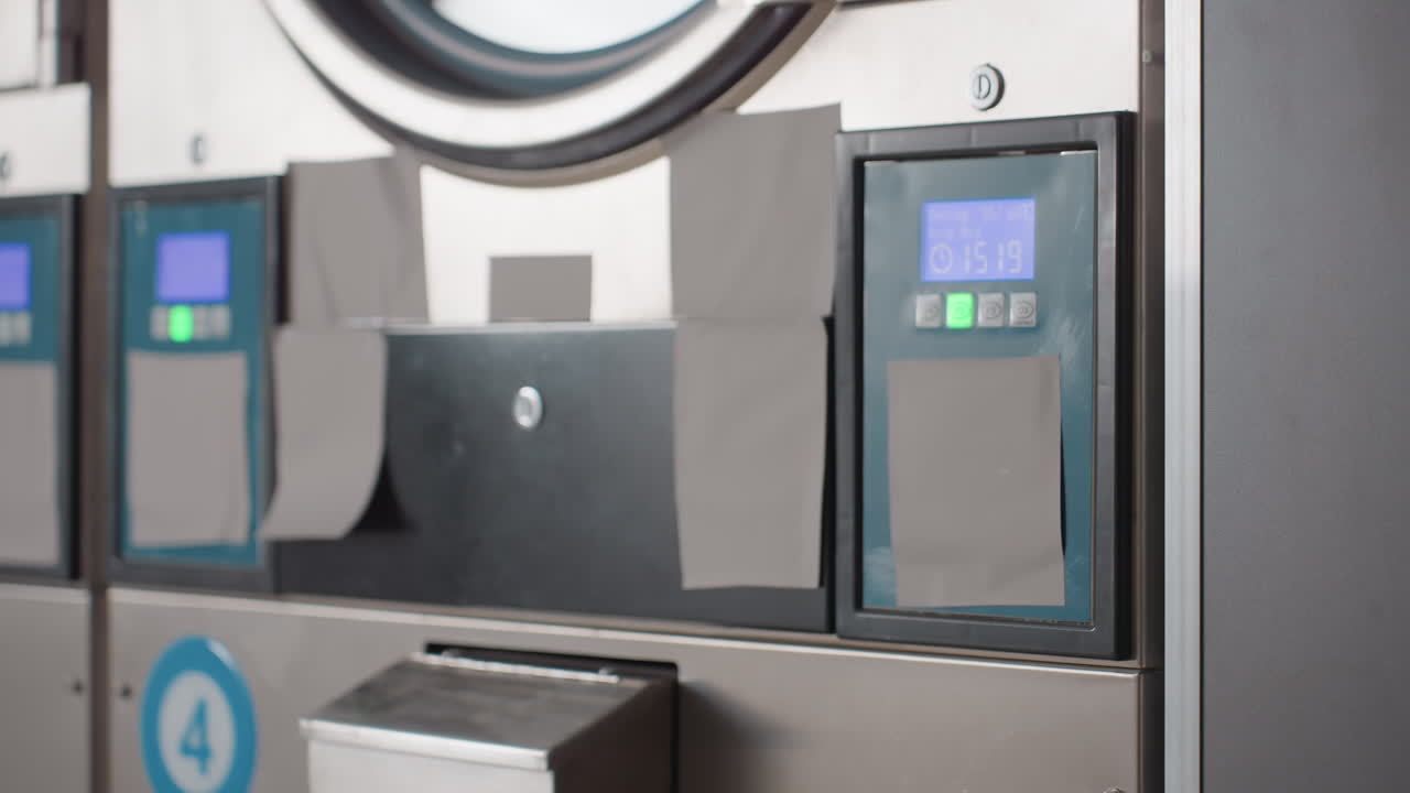 Round view along industrial laundry machines with stainless fronts, digital control panels glowing green, drawers and doors aligned, commercial facility equipment running, clean interior