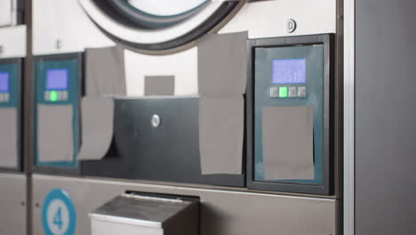 round view along industrial laundry machines with stainless fronts, digital control panels glowing green, drawers and doors aligned, commercial facility equipment running, clean interior