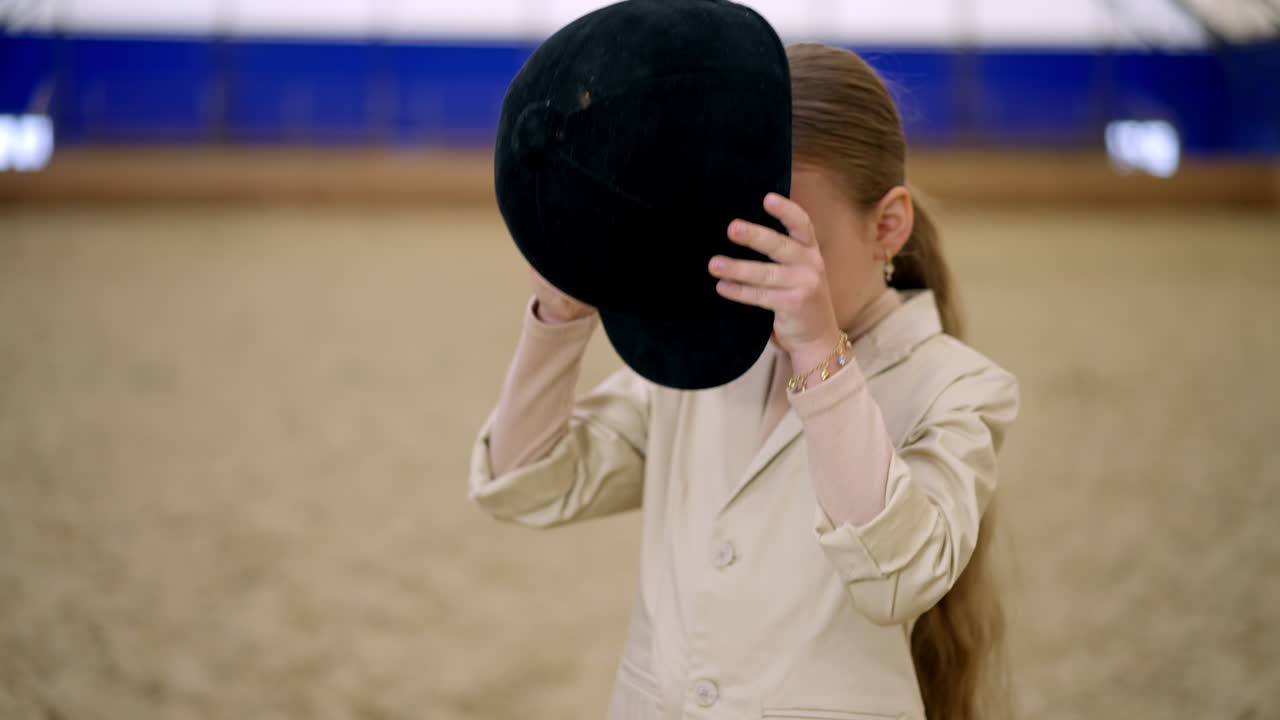 Beautiful young girl in beige jacket puts on a black jockey hat on her head. Lovely little horse rider in riding hall. Close up.