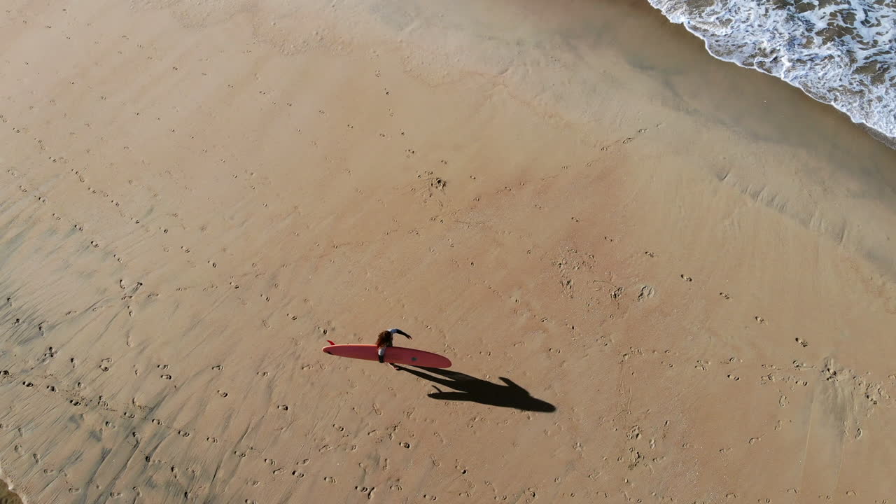 drone aéreo de 4k, surfista jo snider en la playa caminando con una larga tabla de surf al amanecer con largas sombras preparándose para surfear en el muelle de huntington beach, sur de california, océano pacífico