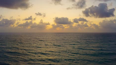 Aerial-drone-shot-slowly-rising-above-the-beach-near-Palm-Beach,-Florida-as-the-waves-crash-and-the-sunrise-begins-to-peek-through-the-clouds