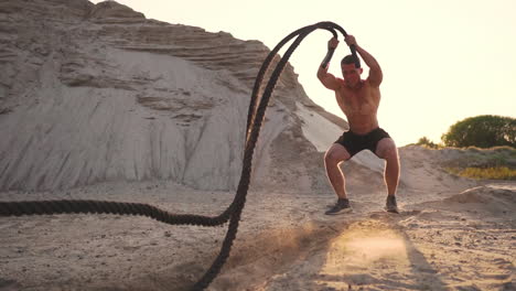 male athlete push-ups on the beach and hits the ground with a rope, circular training in the sun on a sandy beach.