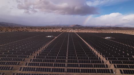 Aerial-Drone-Footage-of-Solar-Panel-Field-in-Joshua-Tree-National-Park-on-a-Sunny-Day-with-rainbow-in-the-background,-forward-moving-shot