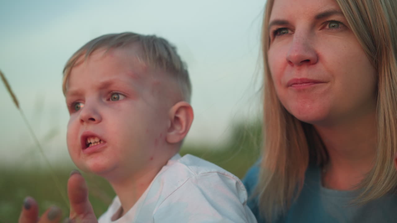 A close-up shot of a mother in a blue gown sitting in a field with her young son in a white shirt on her lap. Both are intently staring at their surroundings, absorbed in the beauty of nature