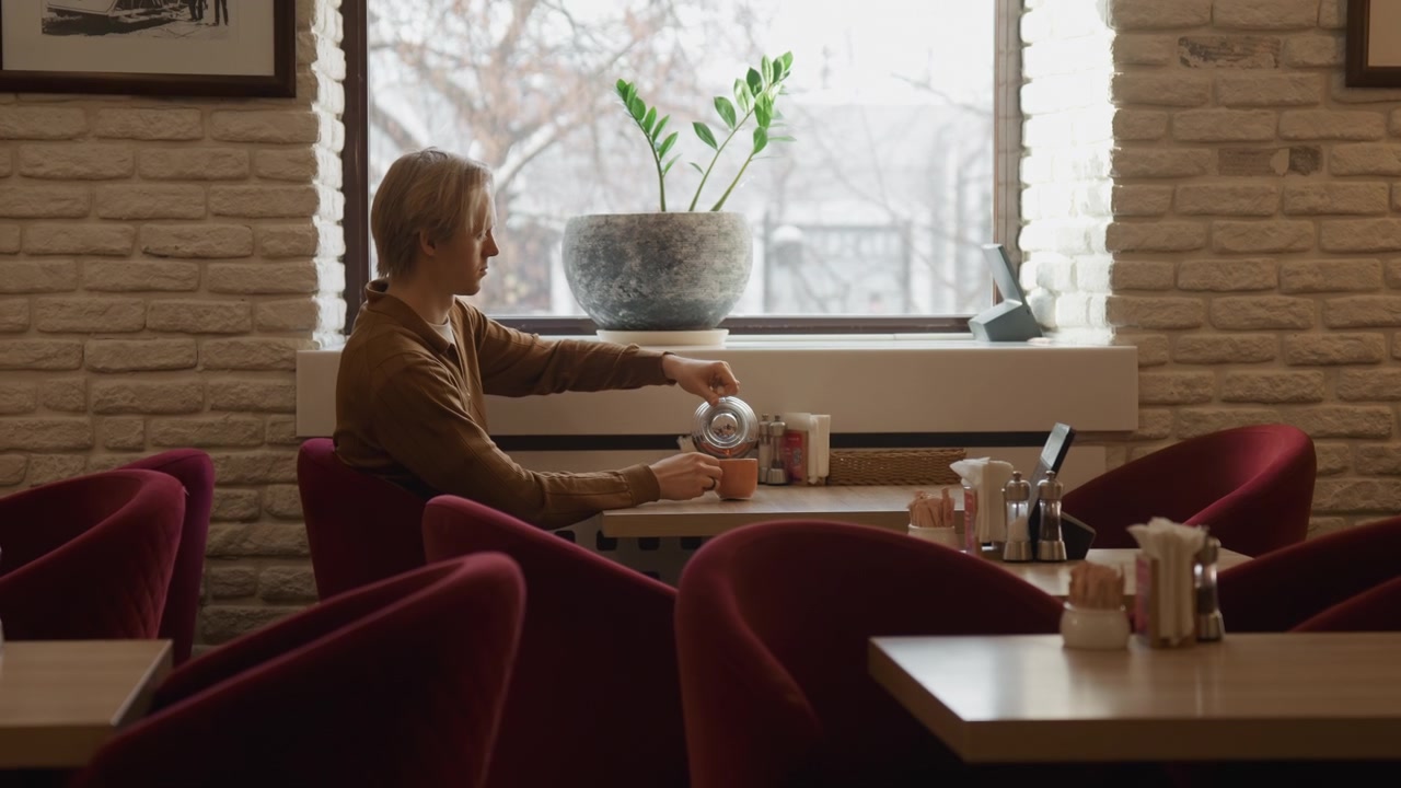 Una donna siede a un tavolo in un caffè al coperto, versando caffè da una caraffa di vetro in una tazza. L'ambiente presenta sedie e tavoli rossi con condimenti sui piani dei tavoli, mentre la luce naturale filtra attraverso grandi finestre.