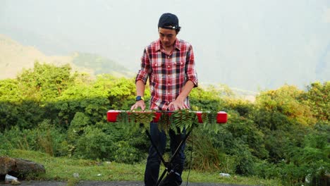 Una-Filmación-Estacionaria-De-Un-Hombre,-Tocando-Su-Teclado-Musical-Electrónico-En-El-Bosque-Cerca-De-Las-Montañas