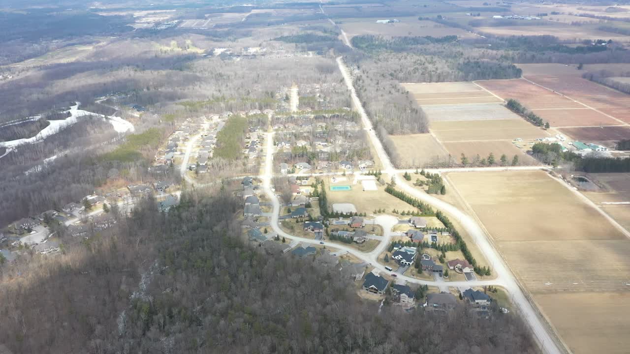 Clip video aerea da drone ad alta quota che sorvola lentamente una comunità suburbana immersa tra una spoglia foresta invernale con chiazze di neve e ampi campi agricoli nella campagna