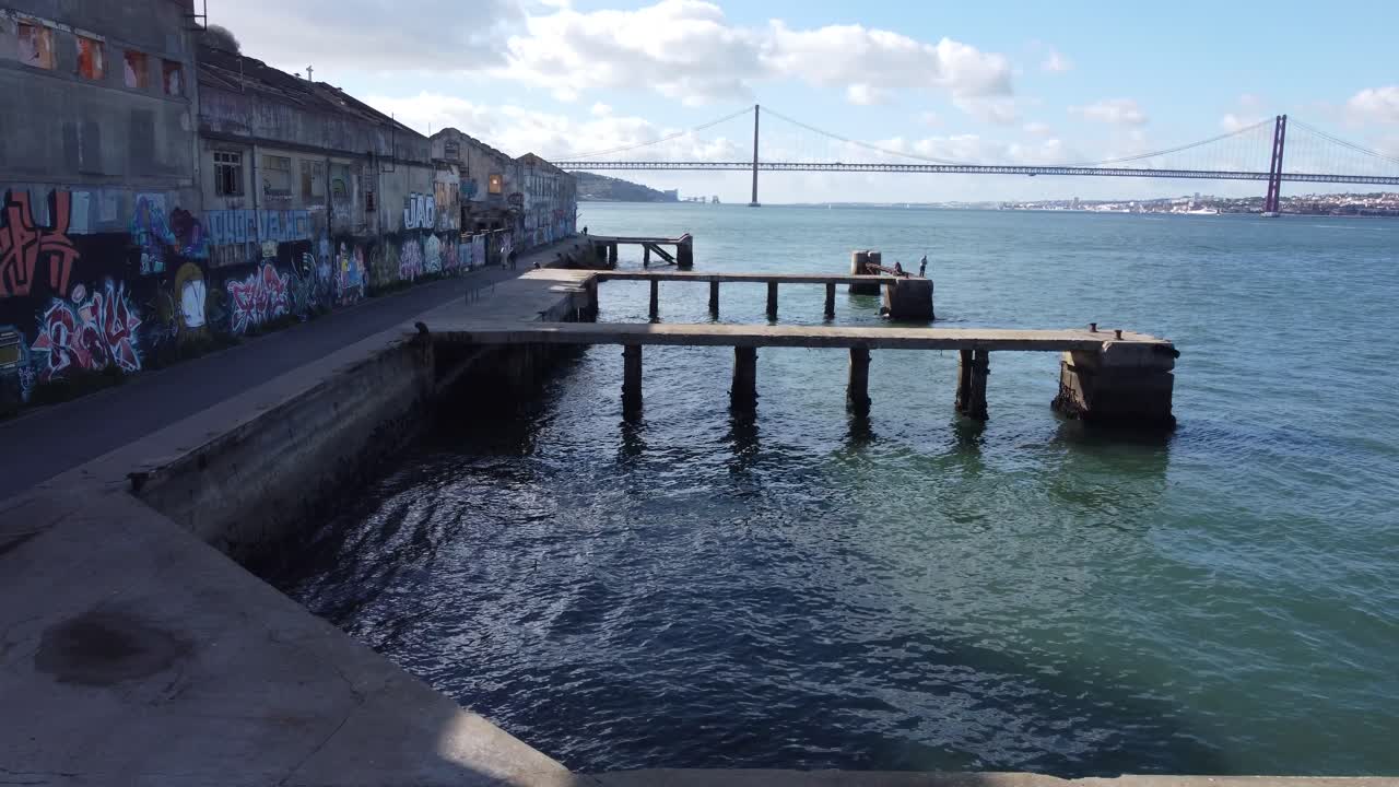 Drone shot flying over some piers next to some abandoned factories, near the river Tejo in Lisbon, ending with an amazing shot of the bridge