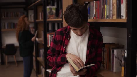 Young-handsome-man-standing-leaning-on-reading-a-book---commuter,-student,-knowledge-concept.-Young-man-in-plaid-shirt-reading-a-book-in-the-college-library