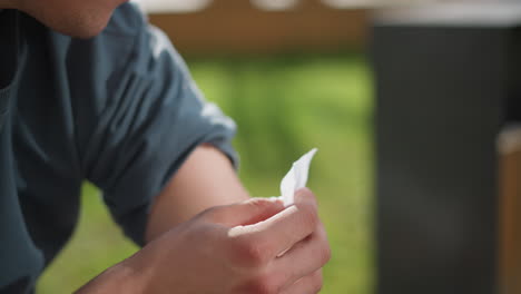 close up view of young white man seated outdoors applying nicotine patch on upper left arm while lifting sleeve, with soft blur background of wooden structure, green grass, and sunlit environment