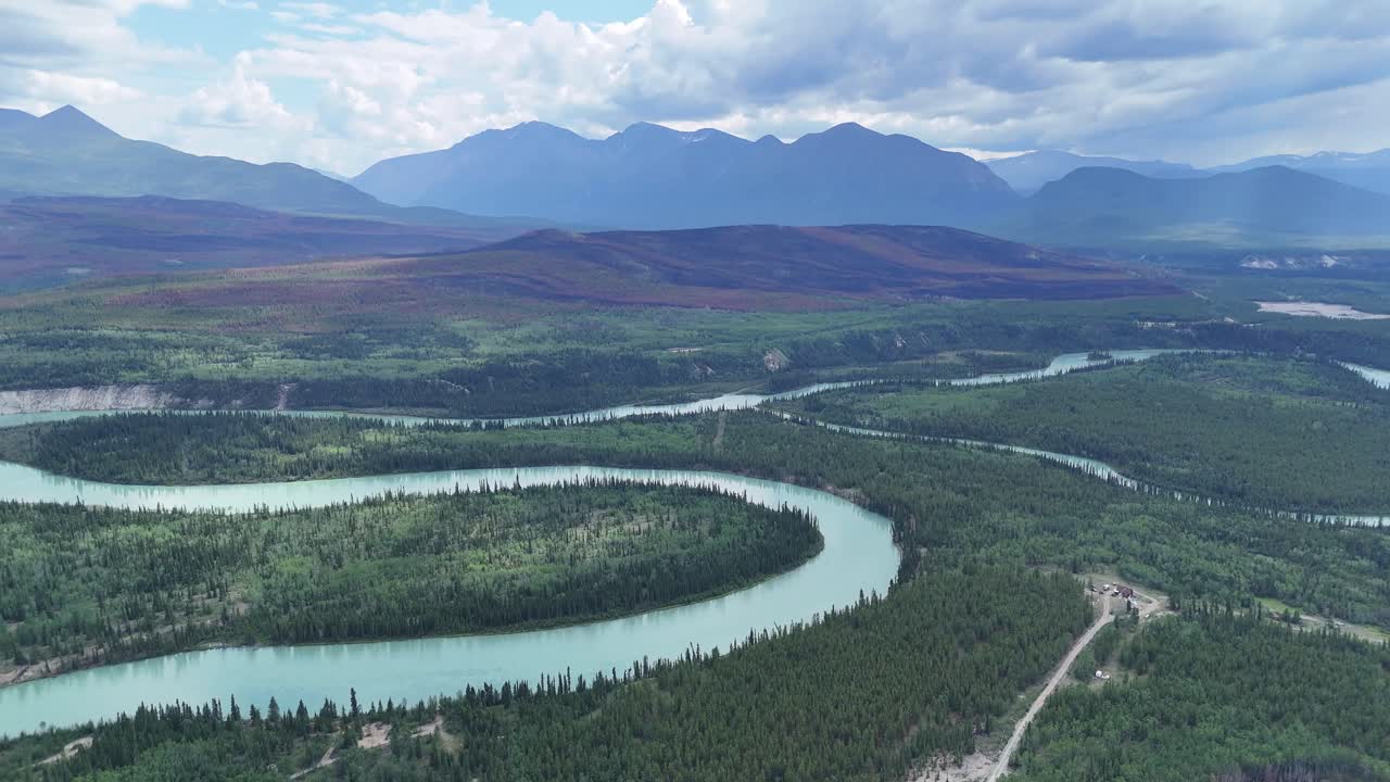 Stunning aerial view captures a winding turquoise river flowing through a vast, verdant forest towards majestic distant mountains under a cloudy sky in Alaska