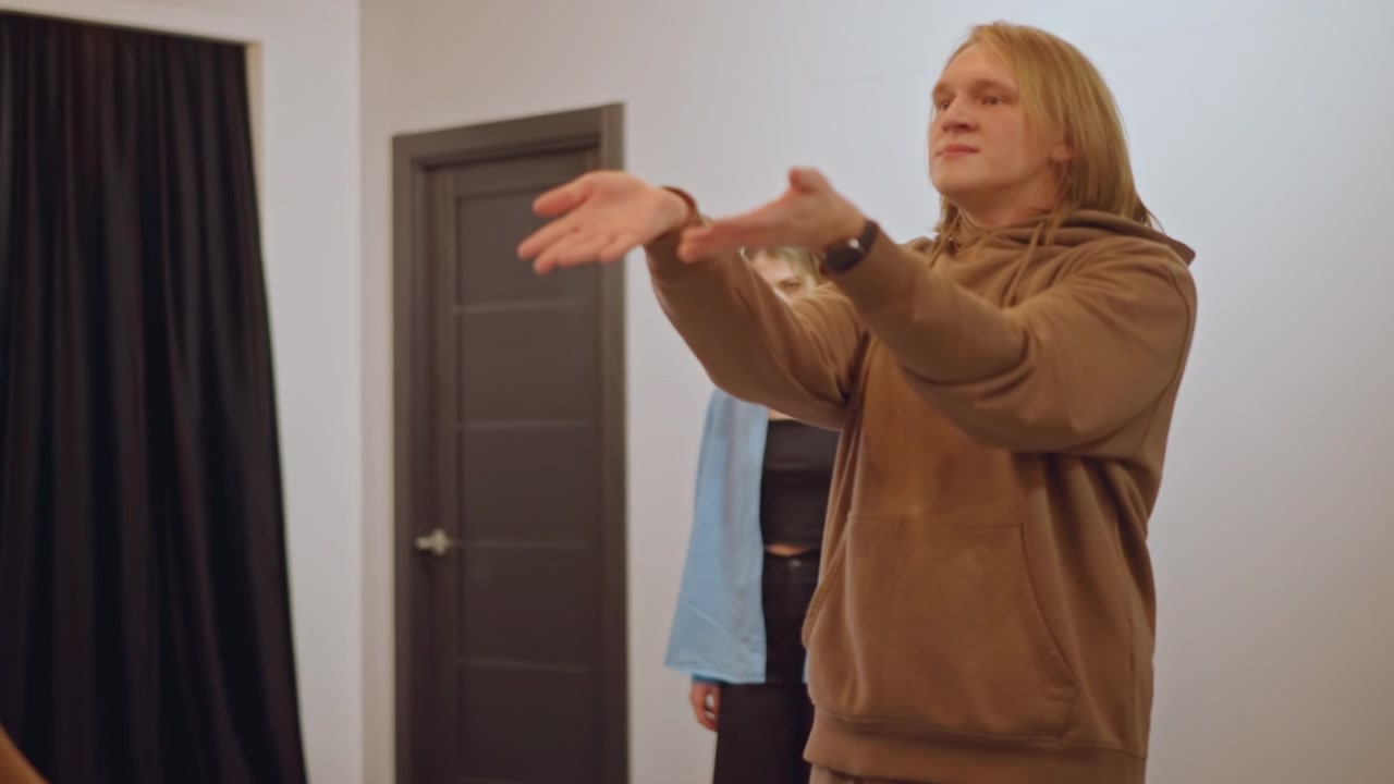 Studentessa bianca in piedi in studio, che osserva il riscaldamento di gruppo mentre l'allenatore gesticola, compagni di classe che praticano il tempo e il ritmo, atmosfera da scuola di musica, esercizi vocali e di movimento, energia in classe.