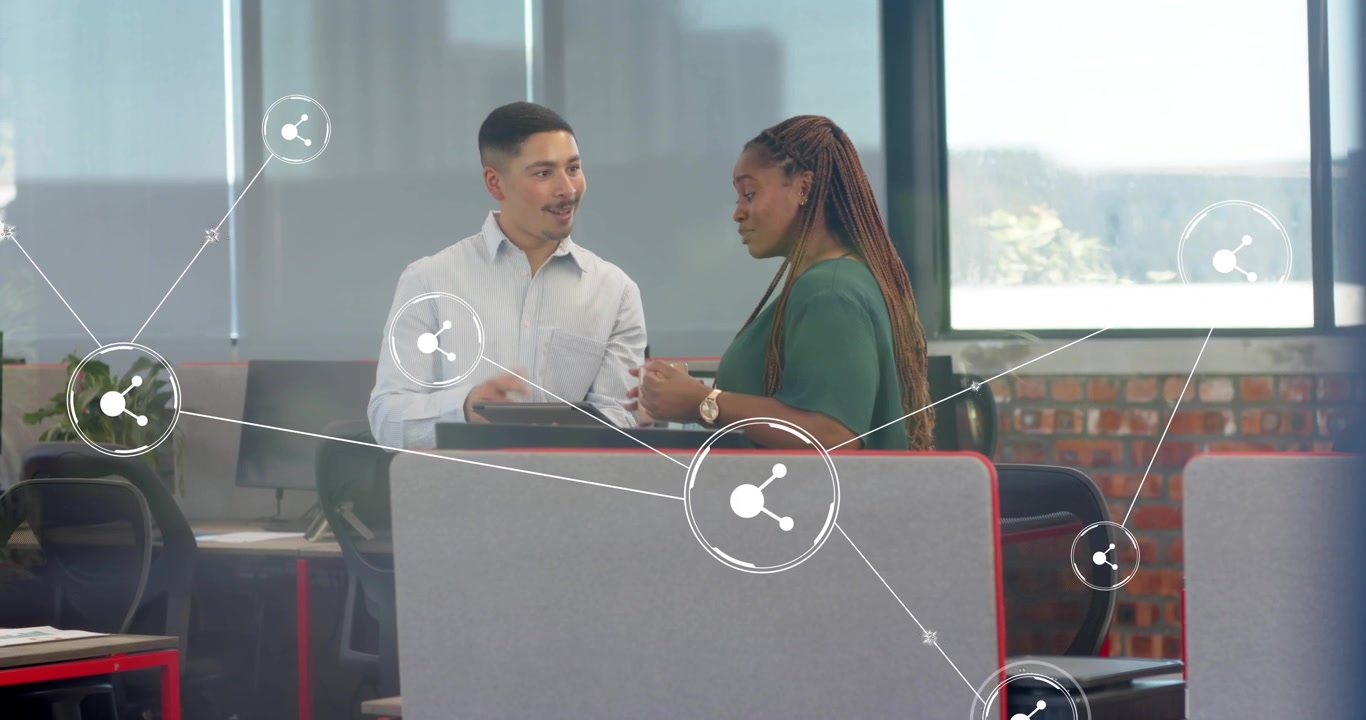 Mid-adult woman approaching desk for business review as man tapping tablet with share icons linking