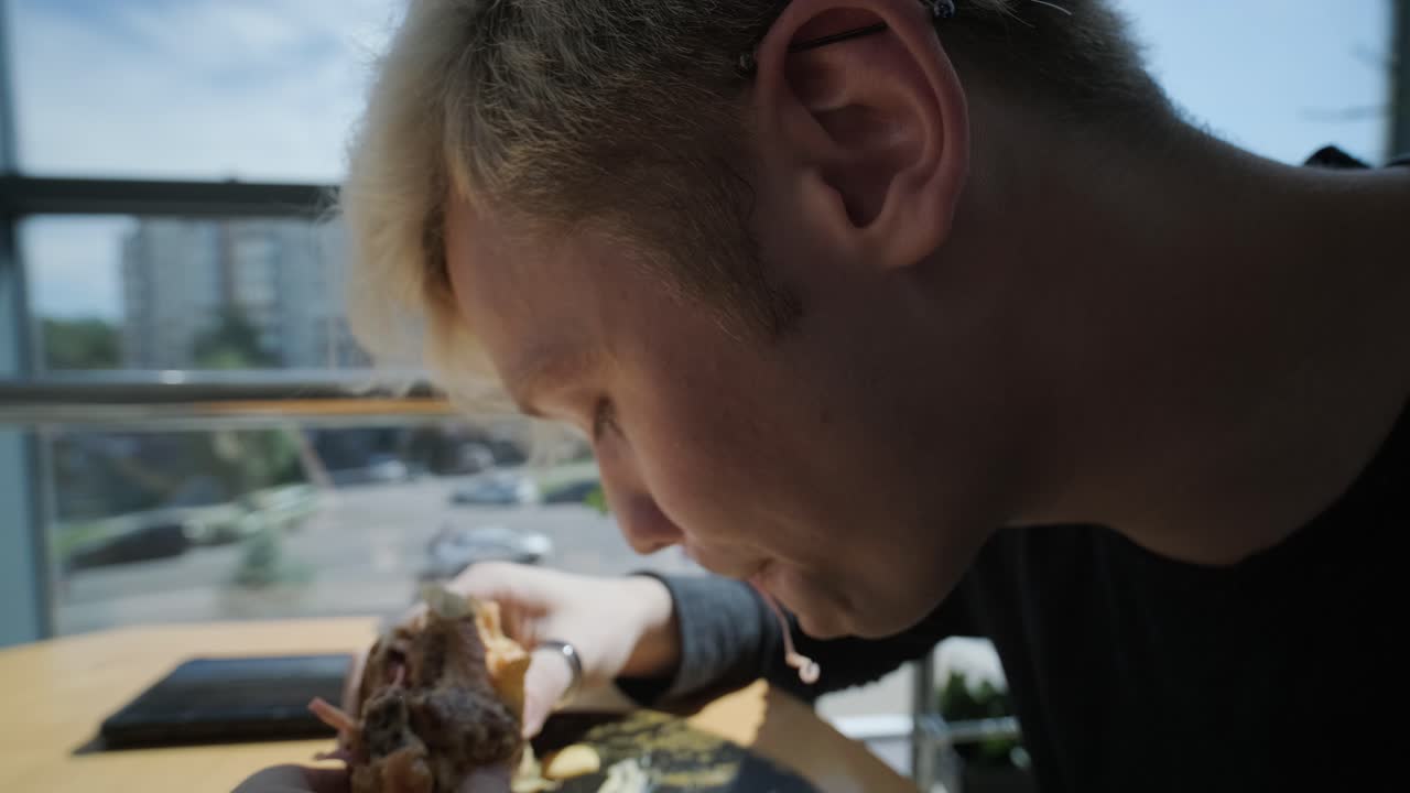 Jovem com cabelo loiro desfrutando de um sanduíche e café gelado em uma mesa de café perto de uma janela com vista para a cidade, luz natural e plantas em vasos. Cena de refeição relaxada e casual