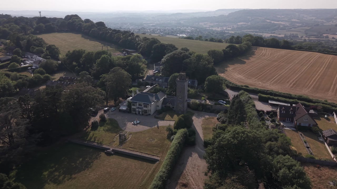 Aerial parallax around a stone tower and country house surrounded by trees, farmland and distant hills.