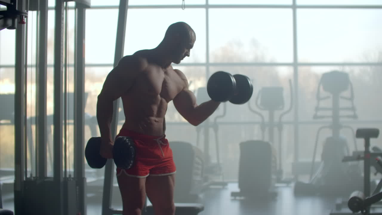 A man with a naked torso and a pumped strong body and relief muscles of the press raises dumbbells against the background of the window training his hands. Strength of will and stubborn character.