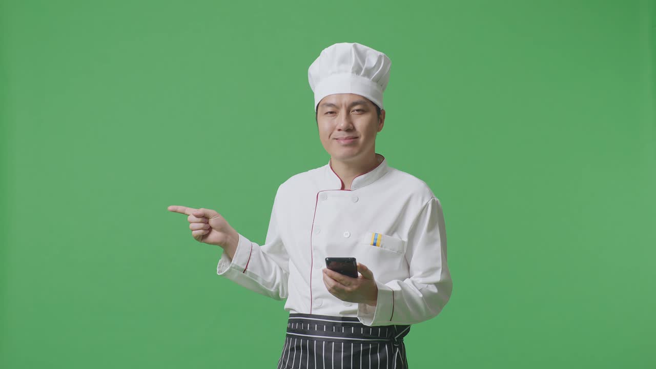 녹색 스크린 배경 스튜디오 에 서 있는 동안 스마트폰 을 사용 하고  을 가리키는 아시아 사람 요리사
