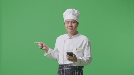 chef asiático usando un teléfono inteligente y señalando hacia un lado mientras está de pie en el estudio de fondo de la pantalla verde