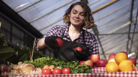 Vista-En-ángulo-Bajo-De-Una-Hermosa-Y-Sonriente-Agricultora-Con-Guantes-Negros-Que-Organiza-Alimentos-Orgánicos-En-Verduras-De-Mercado-Agrícola,-Huevos,-Verduras-De-Pie-En-El-Interior-De-Un-Espacioso-Invernadero