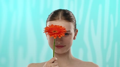 young woman holding gerbera flower with gentle smile a young woman with a gentle smile holds a vibrant orange gerbera flower against a serene blue background