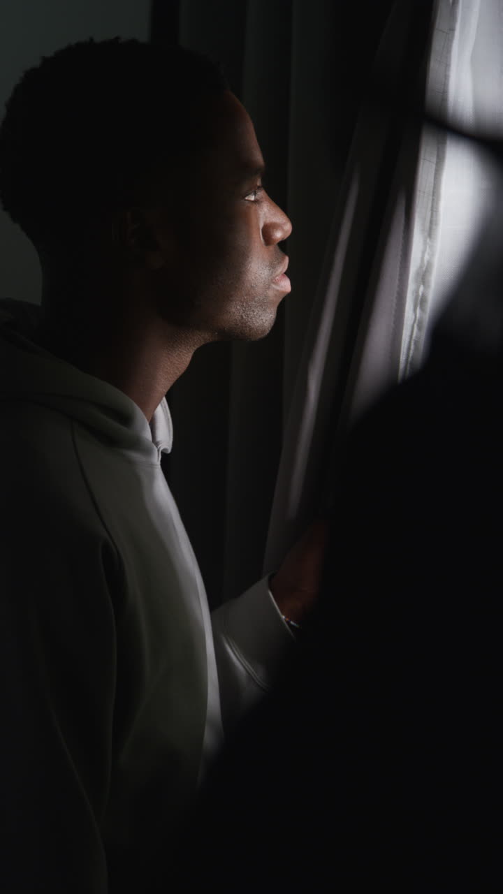 Vertical Video Of Unhappy And Depressed Young Man At Home Looking Out Of Window Suffering With Agoraphobia Or Paranoia
