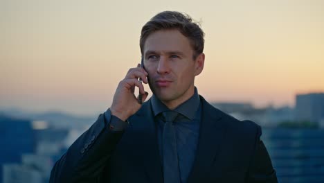 blond caucasian businessman in a suit smiles confidently while talking on his smartphone at an outdoor office terrace with a city skyline at sunset