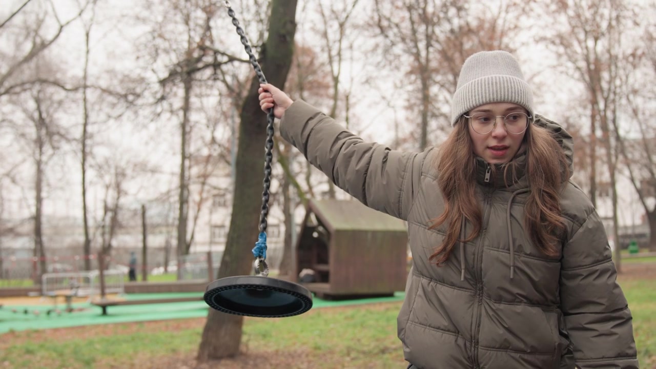 Reflective white girl holding park swing chain, puffer coat and beanie, mossy wooden post and empty playground, contemplative gaze and quiet posture, urban green space in muted winter light
