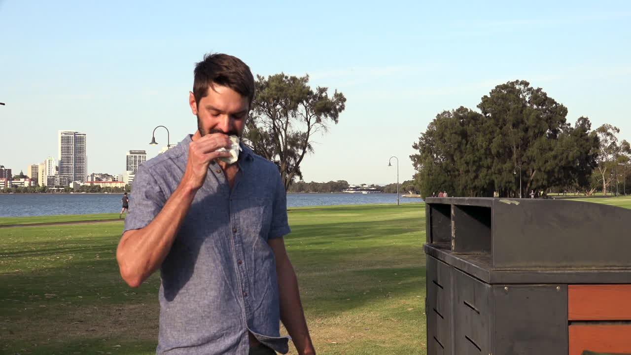 hombre caucásico muerde un sándwich y descarta el resto en un contenedor, en un parque al aire libre