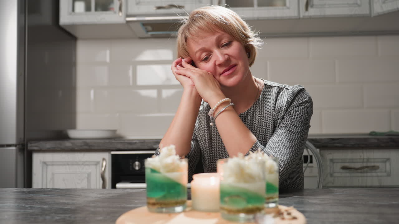 Female chandler supports head with both hands, smiling warmly as she admires beautifully decorated candle glasses on table, filled with green and white wax layers and topped with artistic designs