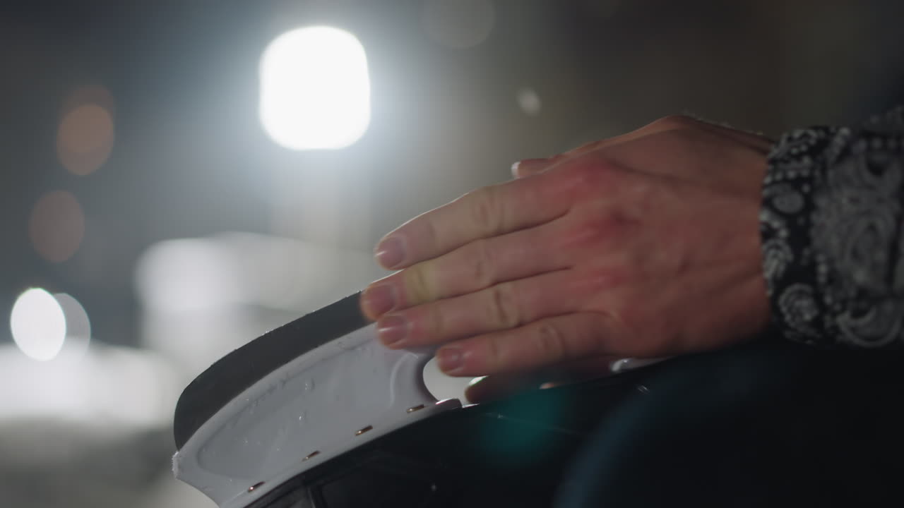 Close up of individual hand gently resting on black skating boot under bright light during winter night with soft background bokeh and subtle lens flare adding atmosphere to the outdoor cold scene