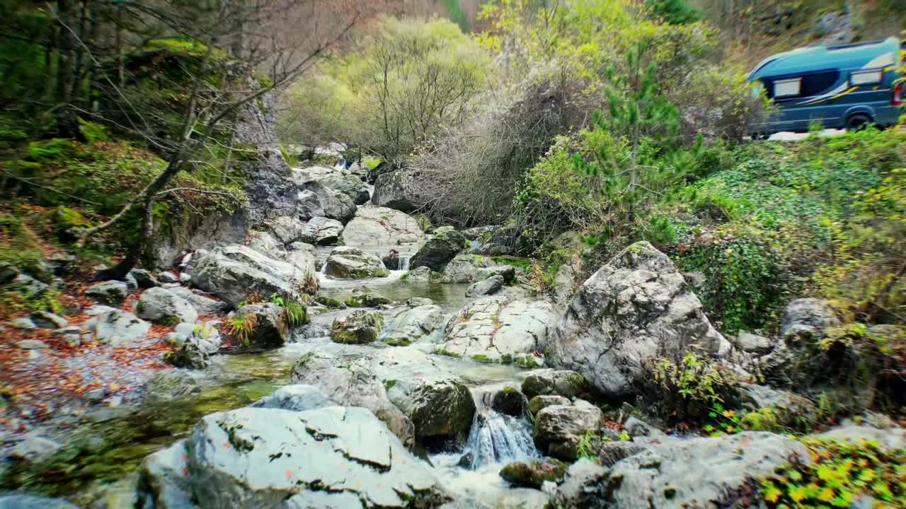 Low drone flight backward over river near parking area at Mount Olympus National Park, Greece, avoiding stones and passing by parked car, capturing natural and human elements